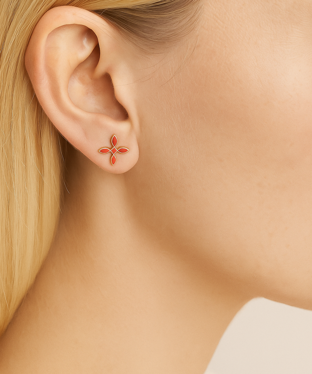 Close-up of an ear wearing a small coral cross earring with a blurred neutral background