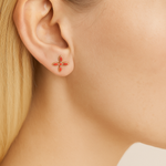 Close-up of an ear wearing a small coral cross earring with a blurred neutral background