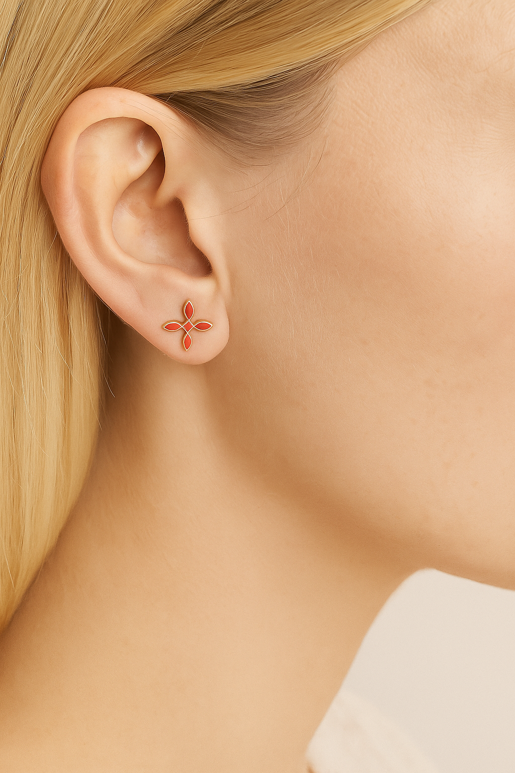 Close-up of an ear wearing a small coral cross earring with a blurred neutral background