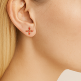 Close-up of an ear wearing a small coral cross earring with a blurred neutral background