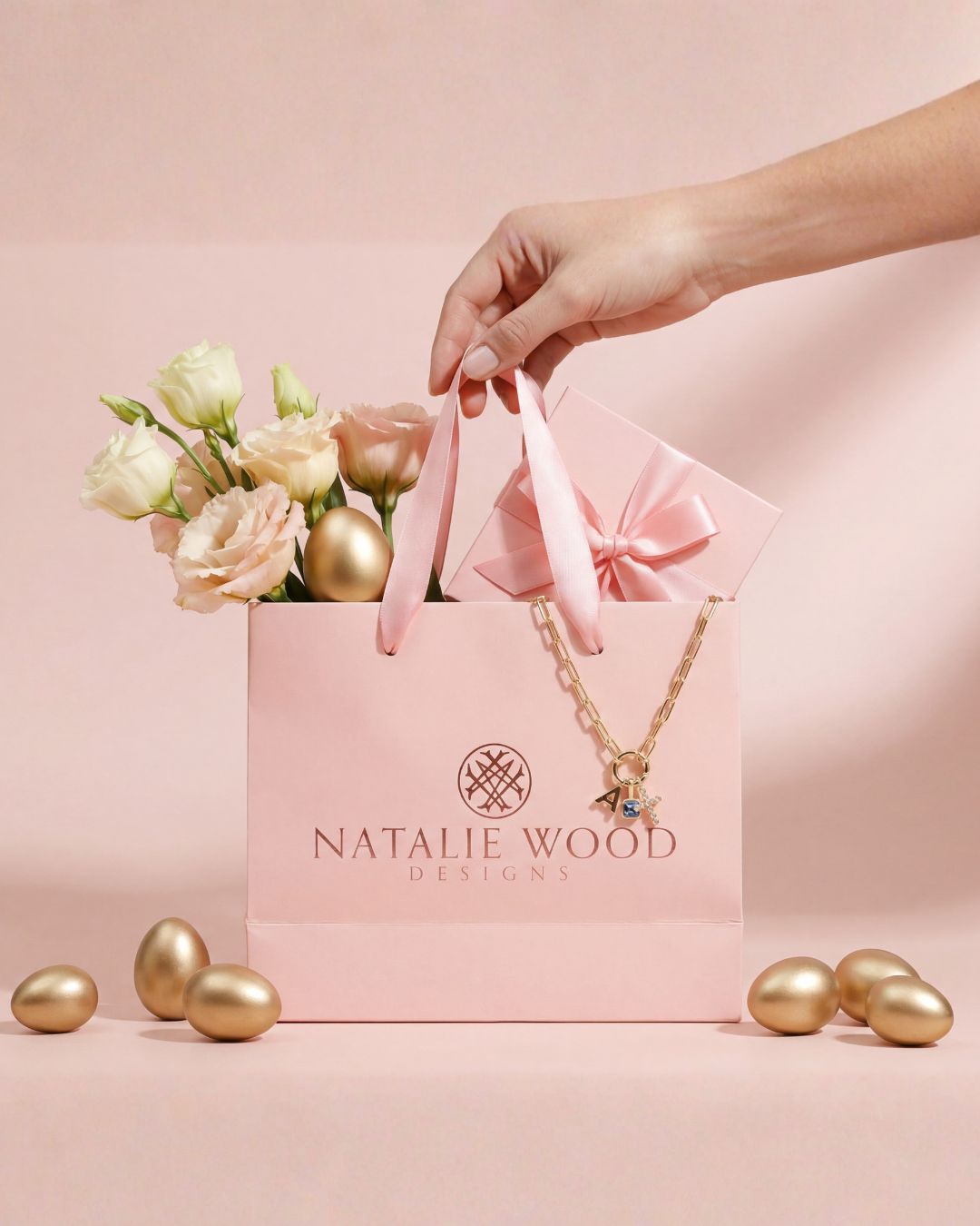 Pink gift bag with 'Natalie Wood Designs' logo, necklace, flowers, and gold eggs on a pink background