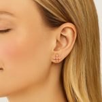 Close-up of a person wearing gold and pink cross earrings with a neutral background